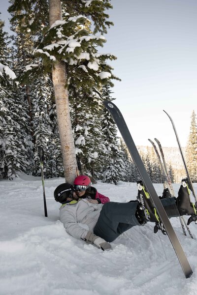 Playful and romantic moment of a couple lying in the snow after skiing in Vail, Colorado, surrounded by snow-covered trees and breathtaking mountain scenery. This adventure elopement captures the fun, intimacy, and spontaneity of a winter wedding experience in the Rocky Mountains.
