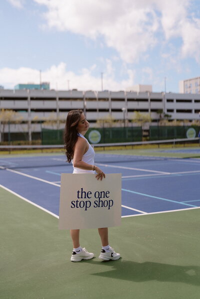 Woman on tennis court holding sign saying the one stop shop