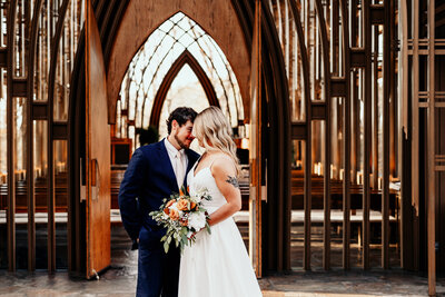 The bride and groom celebrate their marriage by sharing a longing glance and a tight embrace in front of the glass chapel