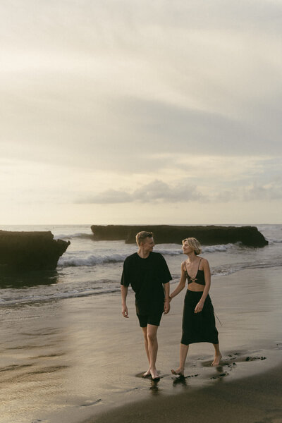 Twee verliefde mensen kijken naar elkaar terwijl ze op een strand van Bali lopen met een ondergaande zon. Vastgelegd door Sharon Kastelijns Photography.