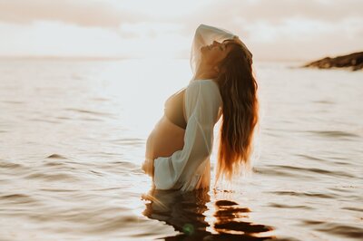 Pregnant woman with a black bikini, with long brown hair stands in the ocean with one hand on her belly and one hand in her hair as she looks up at the sky. The sun is setting behind her