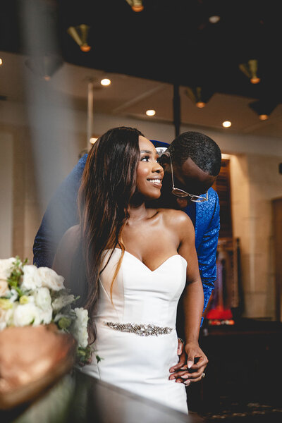 A bride in a strapless white gown smiles while her groom leans in from behind during their wedding ceremony. The couple stands indoors with soft lighting and a floral arrangement in the foreground.