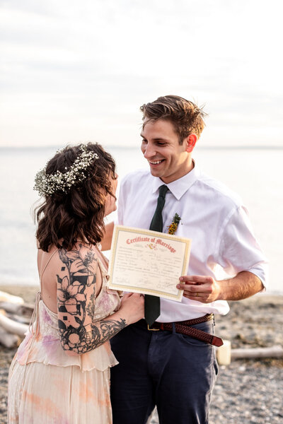 Bride and groom are looking at each other and holding the marriage certificate as they stand along the beach with the water behind them
