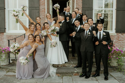 Bride and groom kiss while their wedding party dressed in lavender bridesmaids dresses and black tuxedos cheer around them at Misselwood in Beverly, MA