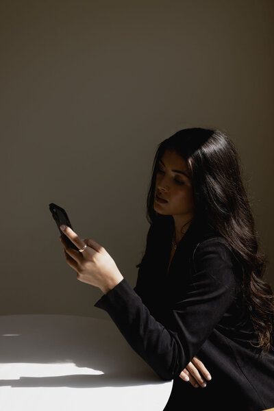 A businesswoman with long black hair in a black suit, seated at a table, focused on her phone, exuding professionalism and poise.