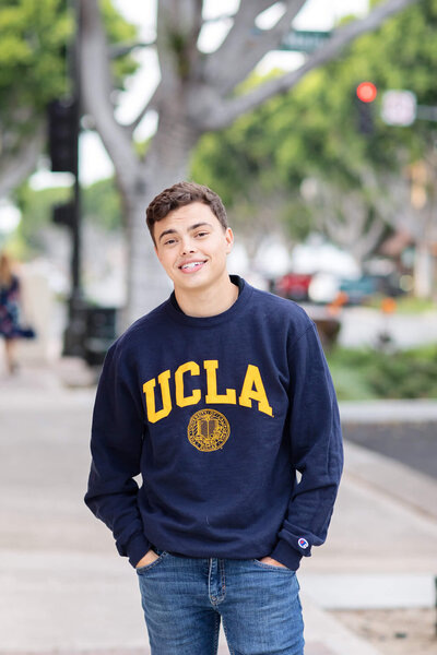 A young man standing on a sidewalk wearing blue jeans and a navy crewneck that reads "UCLA" in yellow lettering, smiling at the camera with brown hair in Old Town Tustin.