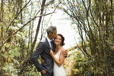 Groom kissing bride’s forehead while embracing outdoors.