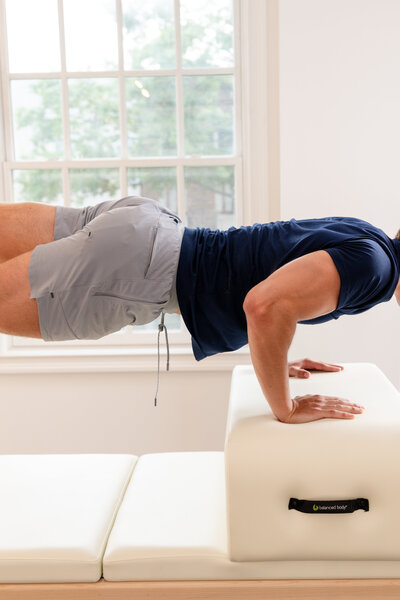 A person in gray shorts and a navy shirt performs a tricep dip, balancing on white foam blocks in a bright room with large windows.