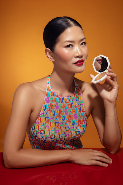 Portrait of Danielle Yu holding a compact mirror, wearing vibrant makeup and a multicolored outfit, captured by Ylva Erevall against a bold orange backdrop.