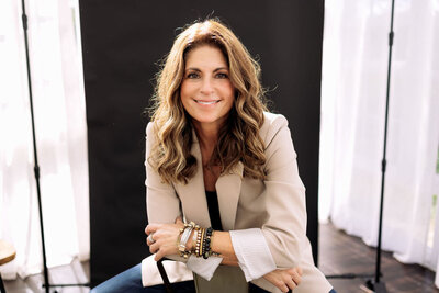 Kerri Link sits in a softly lit studio wearing a beige blazer, smiling warmly with calm confidence and creative energy.