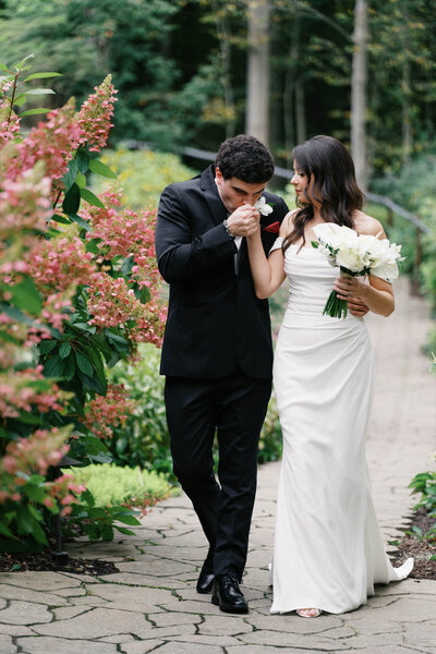 Romantic garden wedding portrait of groom kissing bride’s hand as they walk together, captured by Cleveland OH wedding photographer.