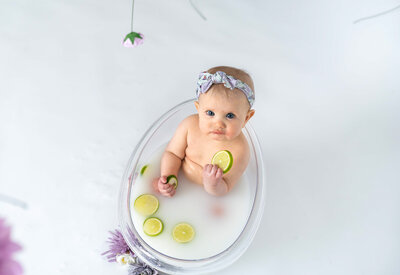 baby girl sitting in clear tub with milkbath  with purple flowers and limes while looking up at camera and holding a lime slice to her mouth