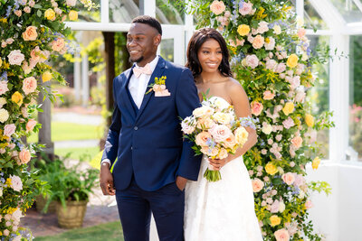 A stunning bride and groom standing before a lush floral archway at an outdoor garden wedding, radiating joy and elegance. The bride holds a vibrant bouquet while the groom looks dapper in a navy suit and pastel bow tie.