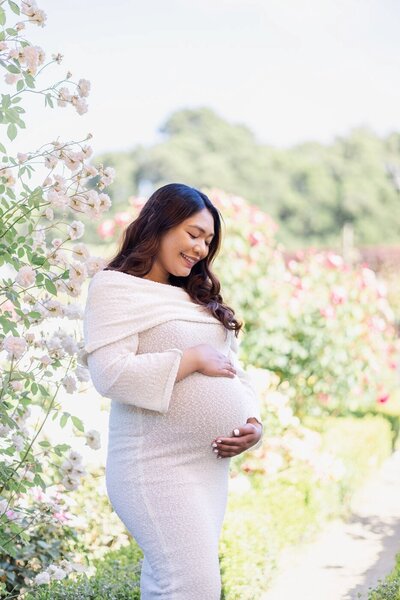 Pregnant woman gazing lovingly at her baby bump amid blooming roses at her maternity photoshoot in Filoli Gardens, Bay Area