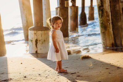 A little girl in a white dress stands near a pier, enjoying a family photo session by the ocean.