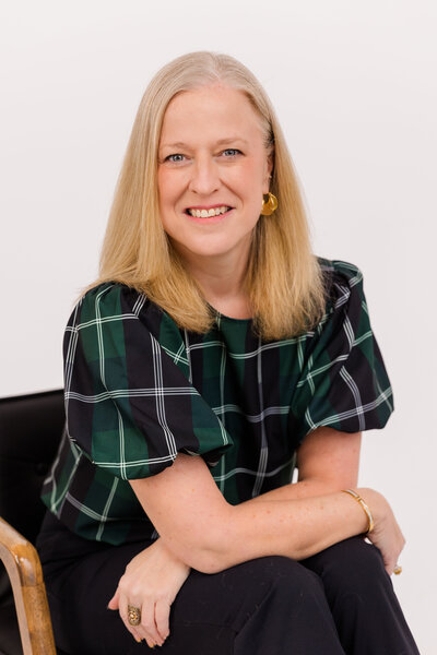 woman with blond hair sitting on a chair in front of white background wearing corporate outfit tartan fabric top and black pant during headshot photoshoot with Laure Photography