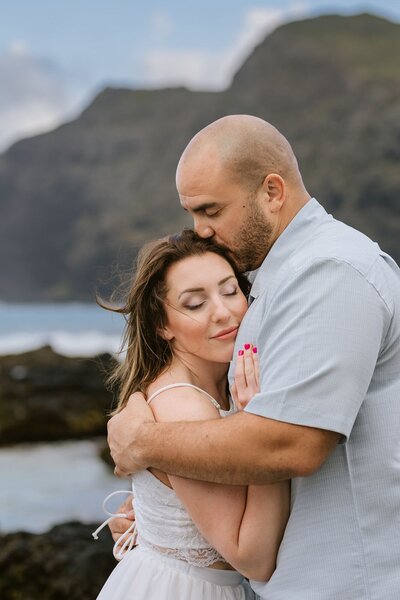 A man holds his wife as he kisses her on the fore head, Rocky Shores, and blue waters behind them.