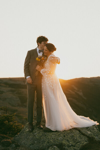Groom in a dark green suit kissing a bride wearing Blundstone shoes on the cheek on the summit of Mount Monroe in the White Mountains of New Hampshire during their elopement.