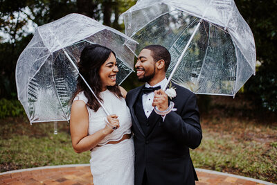 Bride sports a chic two piece set smiles at groom in a tux as they stand outdoors under clear umbrellas.