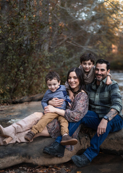 family of four with two young boys poses for outdoor photo sitting on a rock in a low creek shot by juniper &  Oak photo st louis family photographer