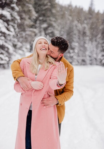 A woman in a pink jacket laughs while being hugged and kissed from behind during their porposal