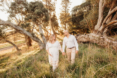 Angela and her husbanding walking along a field of long grass at sunset with maternity photographer michelle kelly