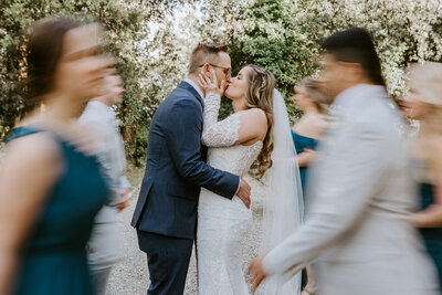 bridge and groom pose kissing while bridal party walks by