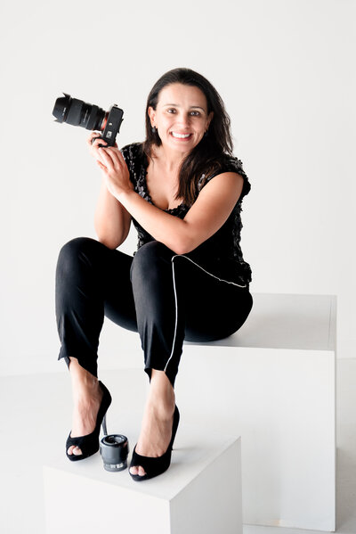 brand photo of a photographer posing with a camera in hand, wearing a black outfit, and seated on white blocks