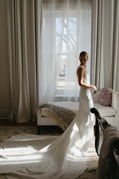 A person in a bridal gown stands near a bright window. Curtains frame the scene, while soft light casts shadows on the floor—captured beautifully by an Ontario destination wedding photographer at Cape Picton. A sofa with a blanket and cushion rests nearby, evoking the essence of wedding film photography.