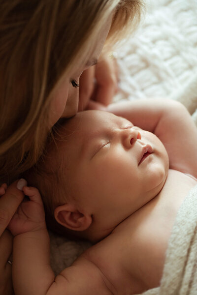 A woman tenderly kisses the forehead of a sleeping baby wrapped in a soft, light-colored blanket. The scene is intimate and serene, capturing a moment of affection often highlighted in Orlando newborn photography sessions.