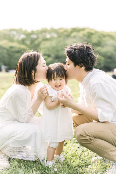 mother and father kissing the head of a cute toddler. Toddler is similing and the backlighting is beautiful in this family portrait. Shot in Tokyo Shibuya by Yukika Ishikawa Photography