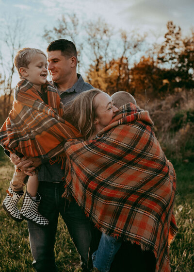 Parents hug on their kids as they are all entangled in a fall colored plaid blanket in the fall at sunset.