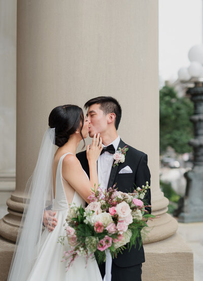 asian couple for a boston city wedding at the fairmont hotel. Bride kisses groom in black tux with stunning florals.
