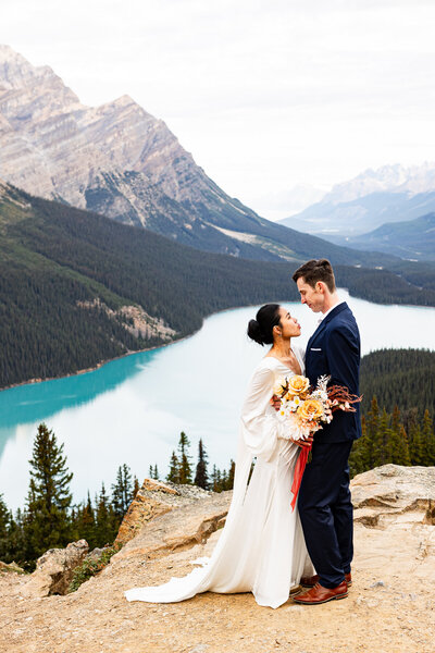 Could in their wedding clothes enjoying a quiet moment on their elopement day. This couple is standing on a mountain overlooking a blue lake. She is wearing a wedding dress and he is wearing a blue suit. they are so happy.