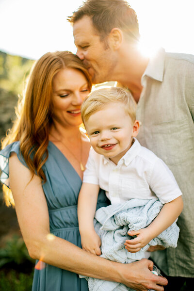 Family Photographer, Mother holding young son with father kissing her forehead