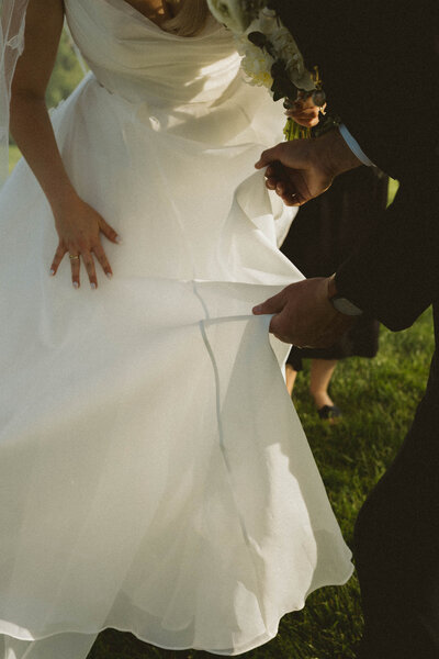 groom looks up to bride smiling as he sits on a grassy hill, bride smiles at camera