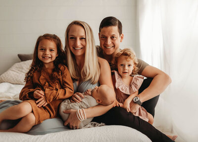 Family of five newborn portrait with mom, dad and big sisters sitting together, smiling at the camera while mom holds newborn baby boy wrapped in neutral tones