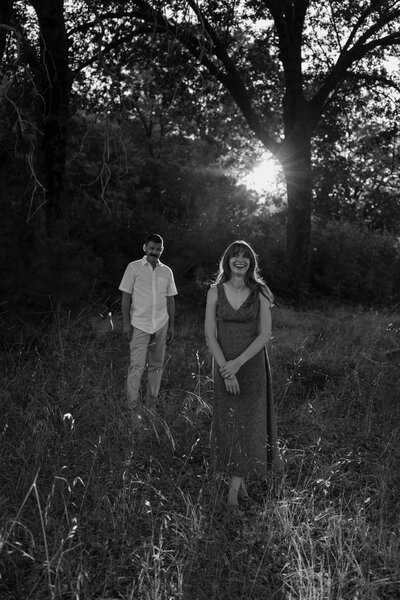 Jordan is standing the the foreground laughing while Wes is standing in the background looking at her, in black and white.