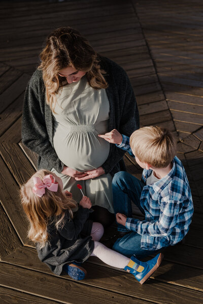 Pregnant mom sitting on her legs kneeling down in green and black white daughter is sitting in front of her looking at belly with lollipop in her hand next to her brother who is in blue plaid drawing with his finger on moms pregnant belly.