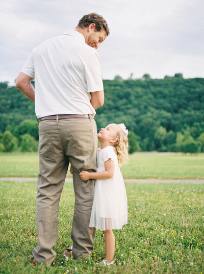 Curly, blonde-haired toddler girl holds onto dad's leg standing in a green field