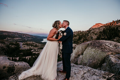 Bride and groom share a kiss while holding their dog during a mountaintop elopement overlooking a lake at sunset near Lake Tahoe.