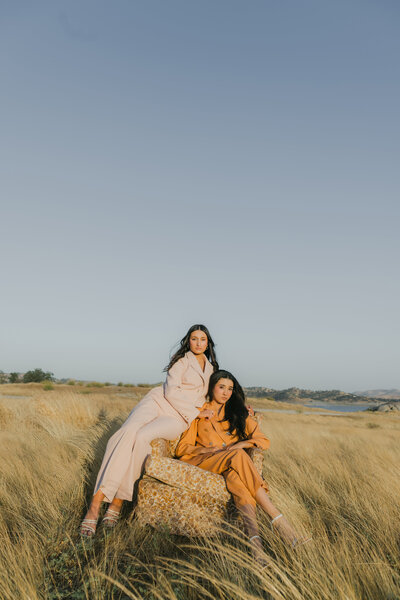 Editorial branding portrait of two sisters in pastel and earth-toned suits walking through a golden field at sunset sitting in a vintage chair, styled for a modern, fashion-forward brand shoot.