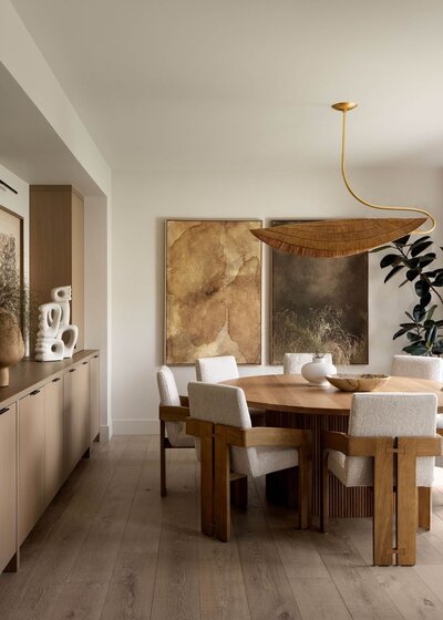 Japandi-style dining space featuring a round fluted dining table, sculptural wood and fabric chairs, oversized rattan pendant, and organic abstract art in earthy tones.