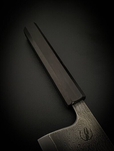 Close-up of the handle of the chef's knife, highlighting its dark color and smooth finish, against a dark background.