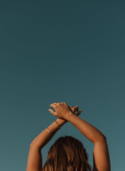 Woman from behind with arms above her head crossed at the wrists, standing in front of blue sky