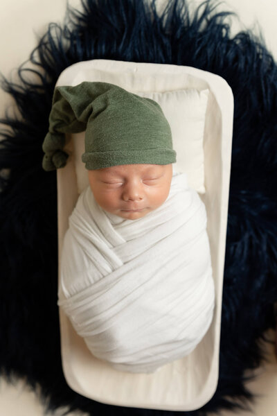 Newborn baby wrapped in a white swaddle wearing a green hat and laying in a white bowl