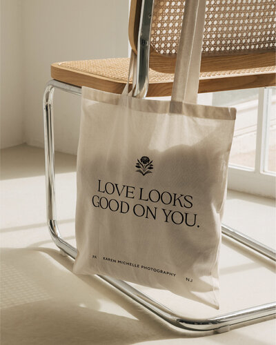 Canvas tote hanging from a cane chair printed with a branded tagline ‘LOVE LOOKS GOOD ON YOU.’ and the floral submark, part of the wedding photographer brand identity collateral.
