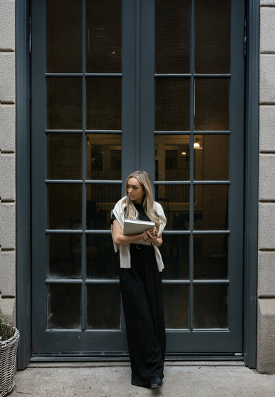 Contemporary branding photo of a woman in a black jumpsuit holding a laptop in front of a dark French-style door — professional, stylish, and captured by Dani Lacey in Greenville SC.