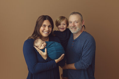 Newborn family photo of mom holding new baby, dad holding toddler son all wearing blue on a tan background.