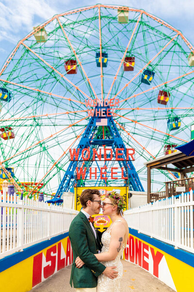 A couple with their arms around each other standing nose to nose in front of a ferris wheel on a pier.